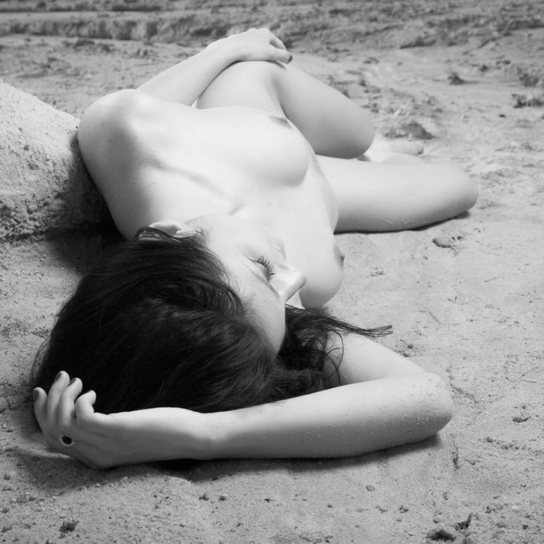 Fekete-fehér érzéki művészi akt homokban fekvő csinos fiatal nővel.